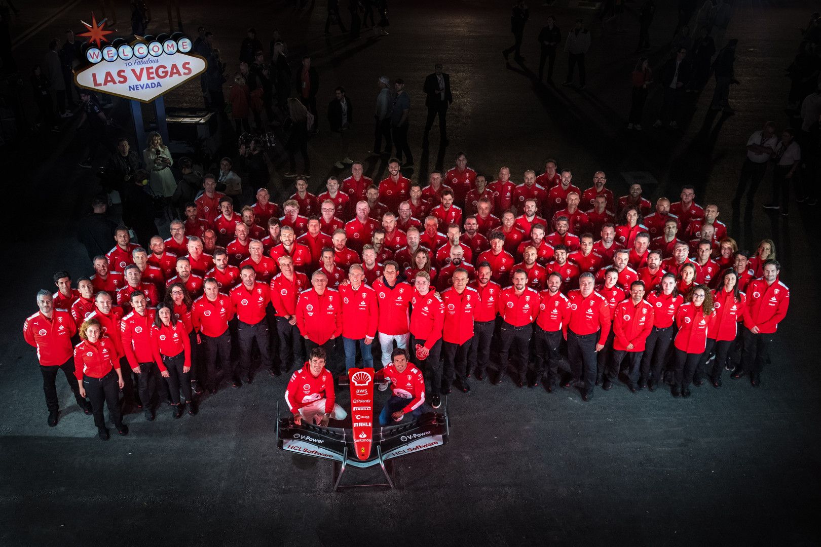 A large team in red stands with a racing car in front of the Welcome to Las Vegas sign.