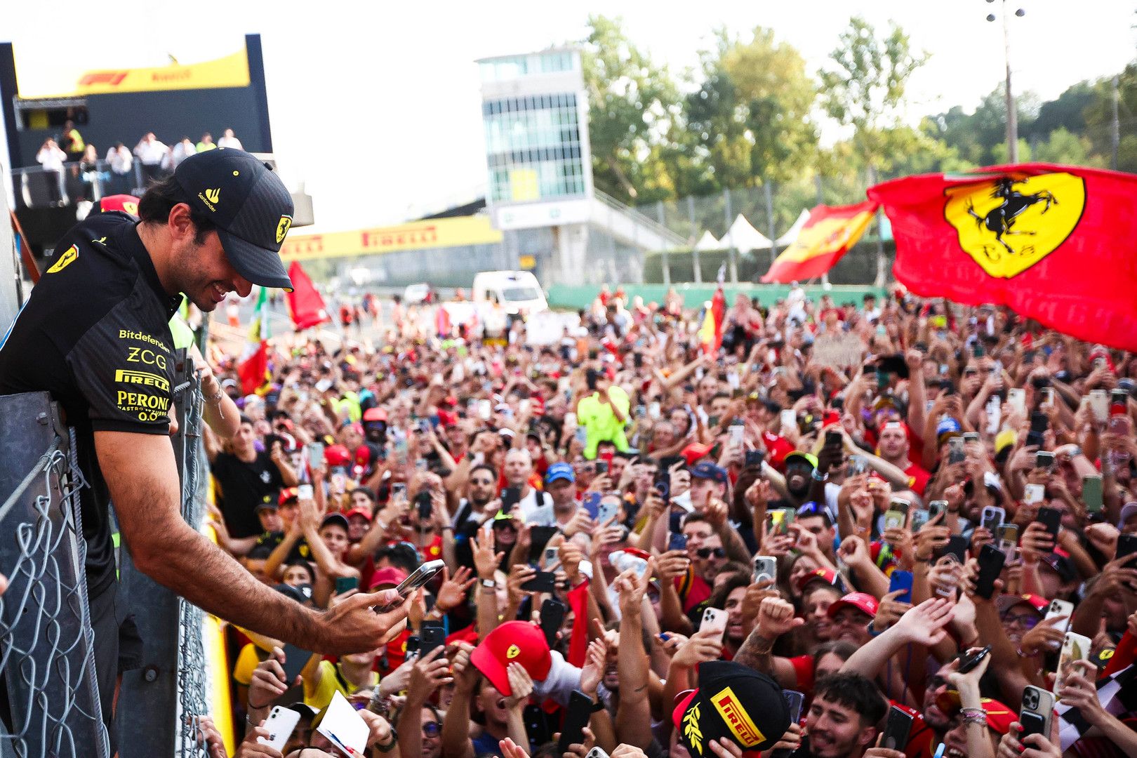Carlos Sainz leans into a cheering crowd of fans holding phones and Ferrari flags.