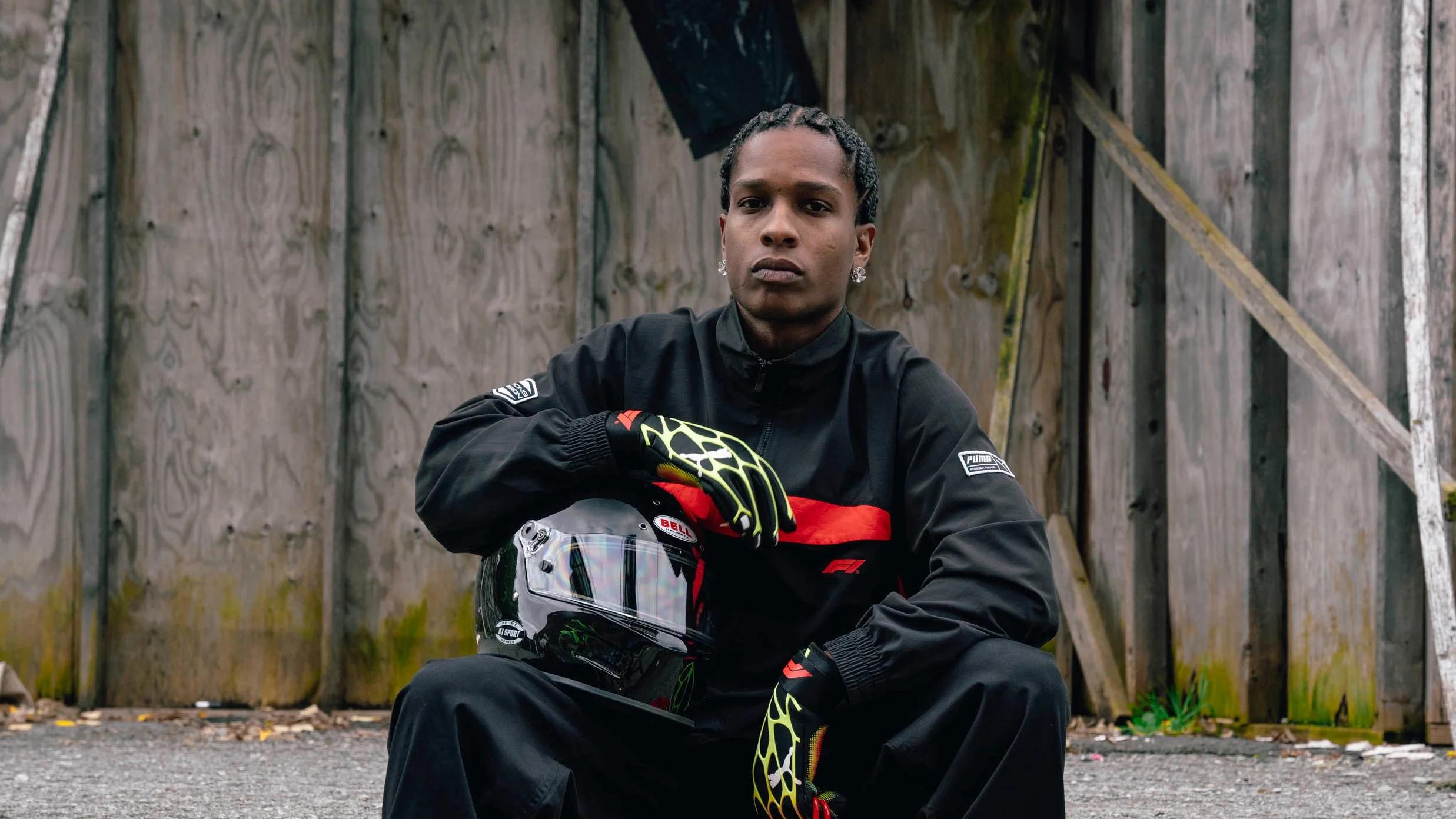 A$AP Rocky in a black racing jacket with a red stripe and yellow gloves, holding a black helmet.