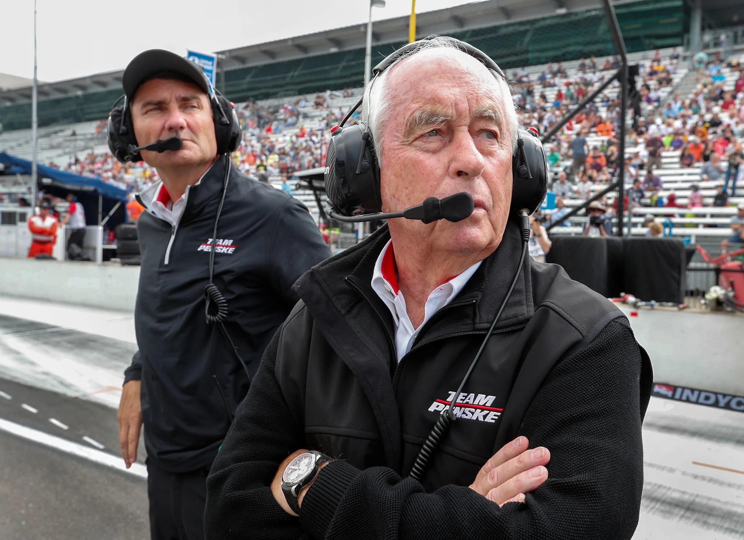 Roger Penske and Tim Cindric, wearing Team Penske jackets and headsets, observe a racetrack.