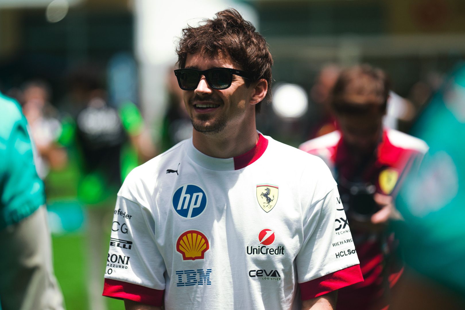 Charles Leclerc smiles, wearing sunglasses and a white Ferrari team shirt with sponsor logos.