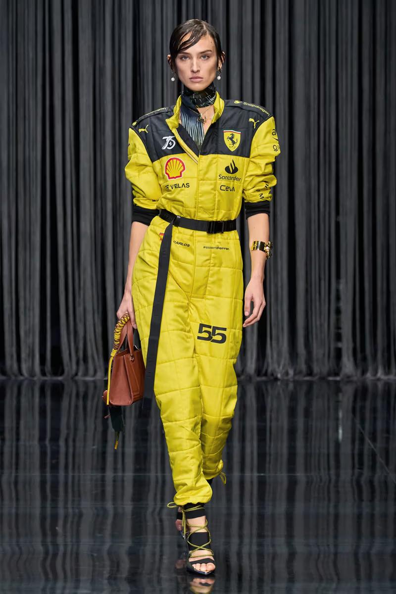 A model on a runway wears a bright yellow racing-style jumpsuit with black details, sponsor logos, the number 55, a brown purse, and black lace-up sandals.