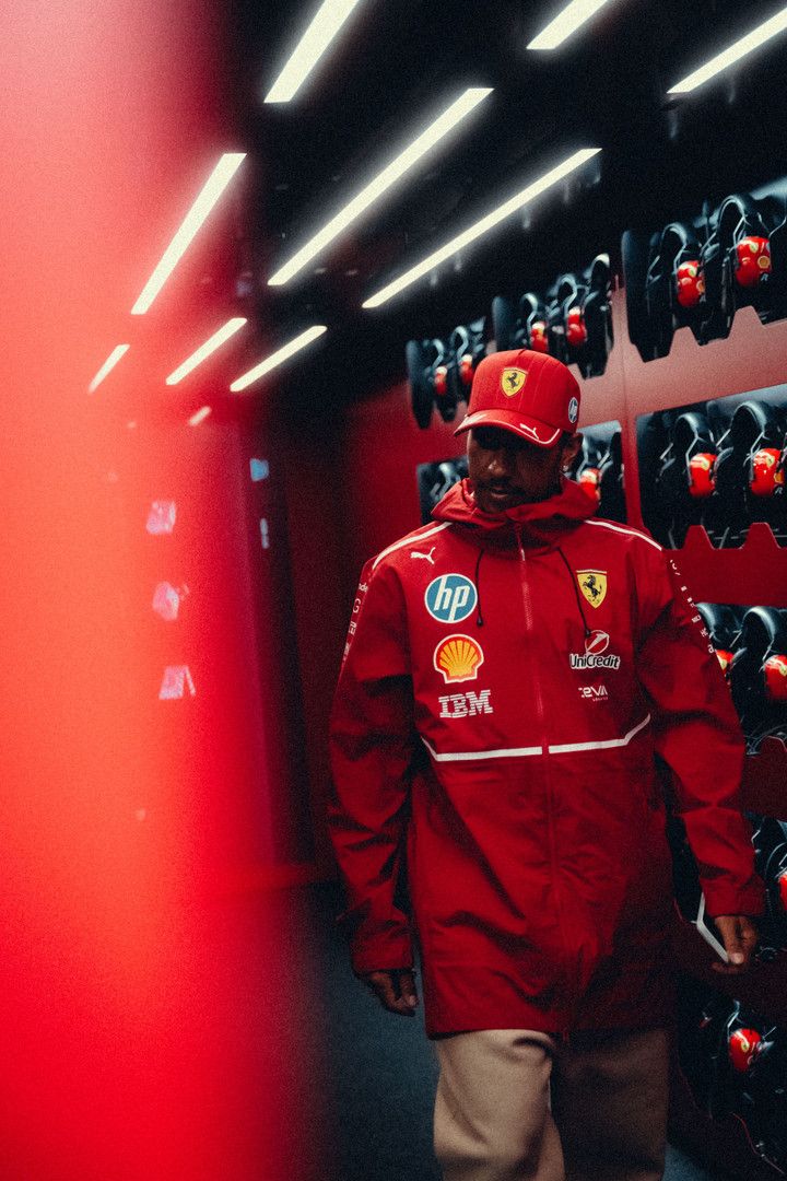 Lewis Hamilton walks through a red-lit corridor wearing a red Ferrari team jacket and cap.
