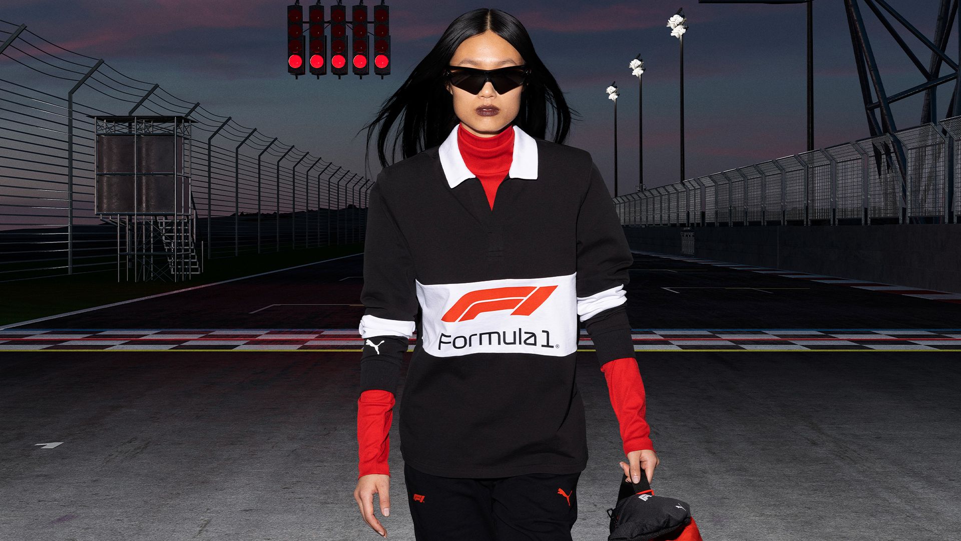 A person wearing a black, white, and red Formula 1 branded outfit and sunglasses walks on a race track with red start lights overhead.