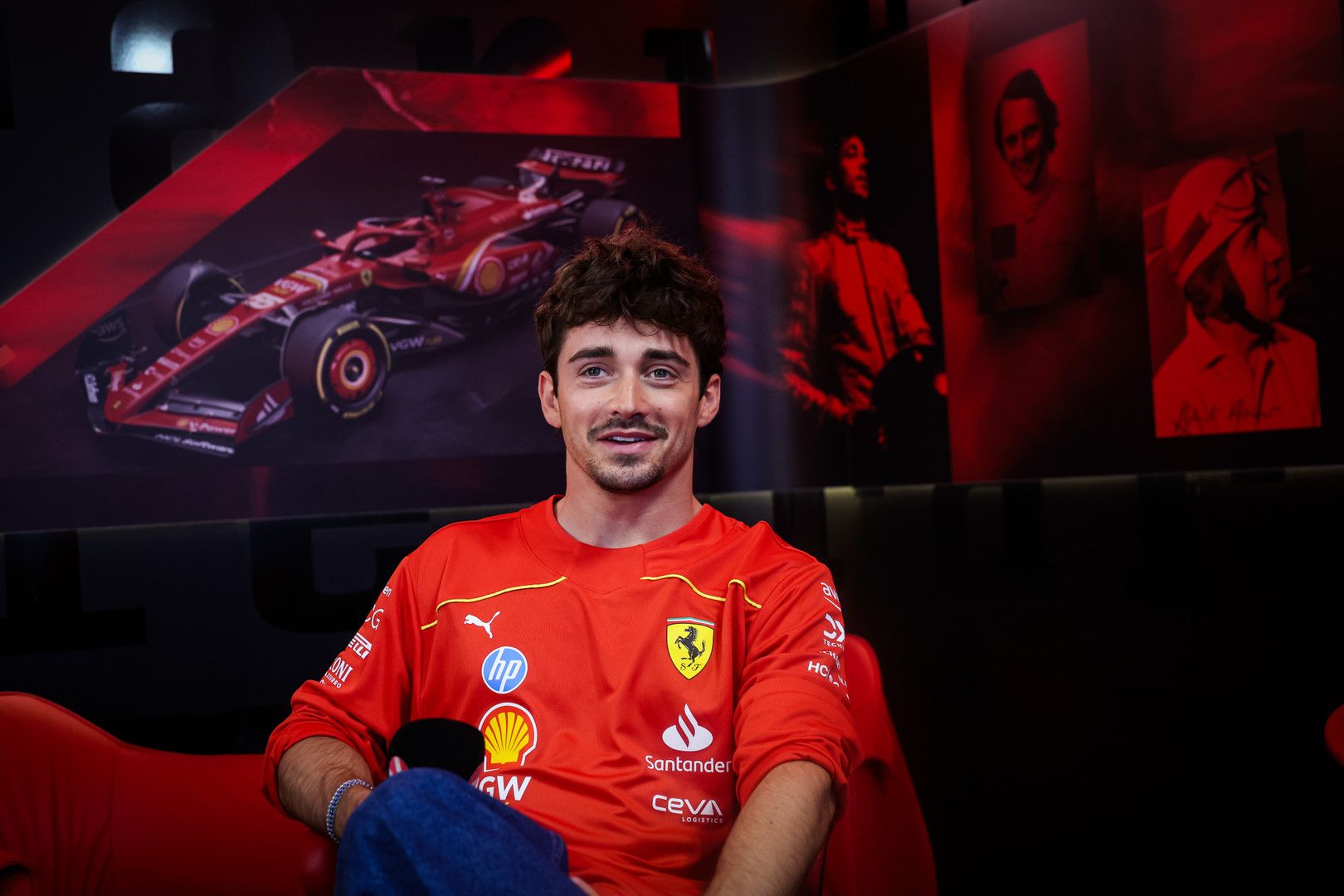 Charles Leclerc smiles in a red Ferrari team shirt with an F1 car and historic racing figures in the background.