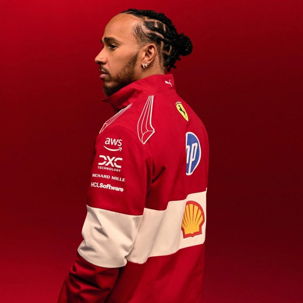 Lewis Hamilton in a red and white racing jacket, looking left in profile against a red background.