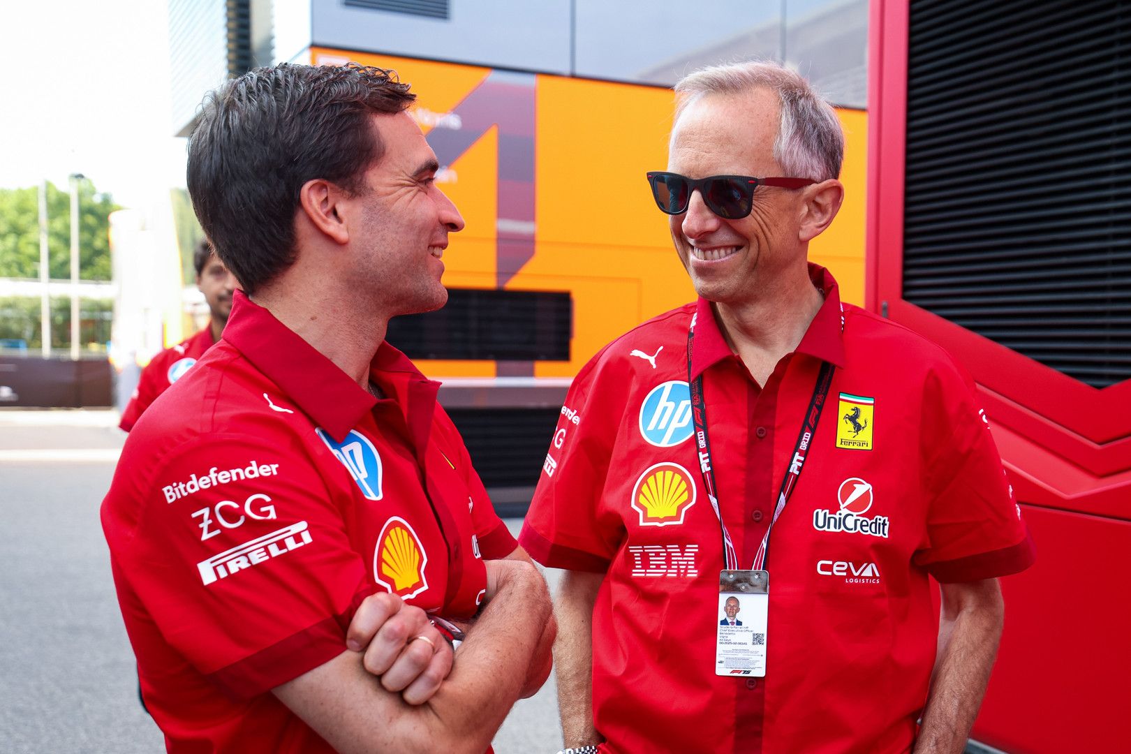 Two men in red Ferrari team shirts smile at each other, one wearing sunglasses.