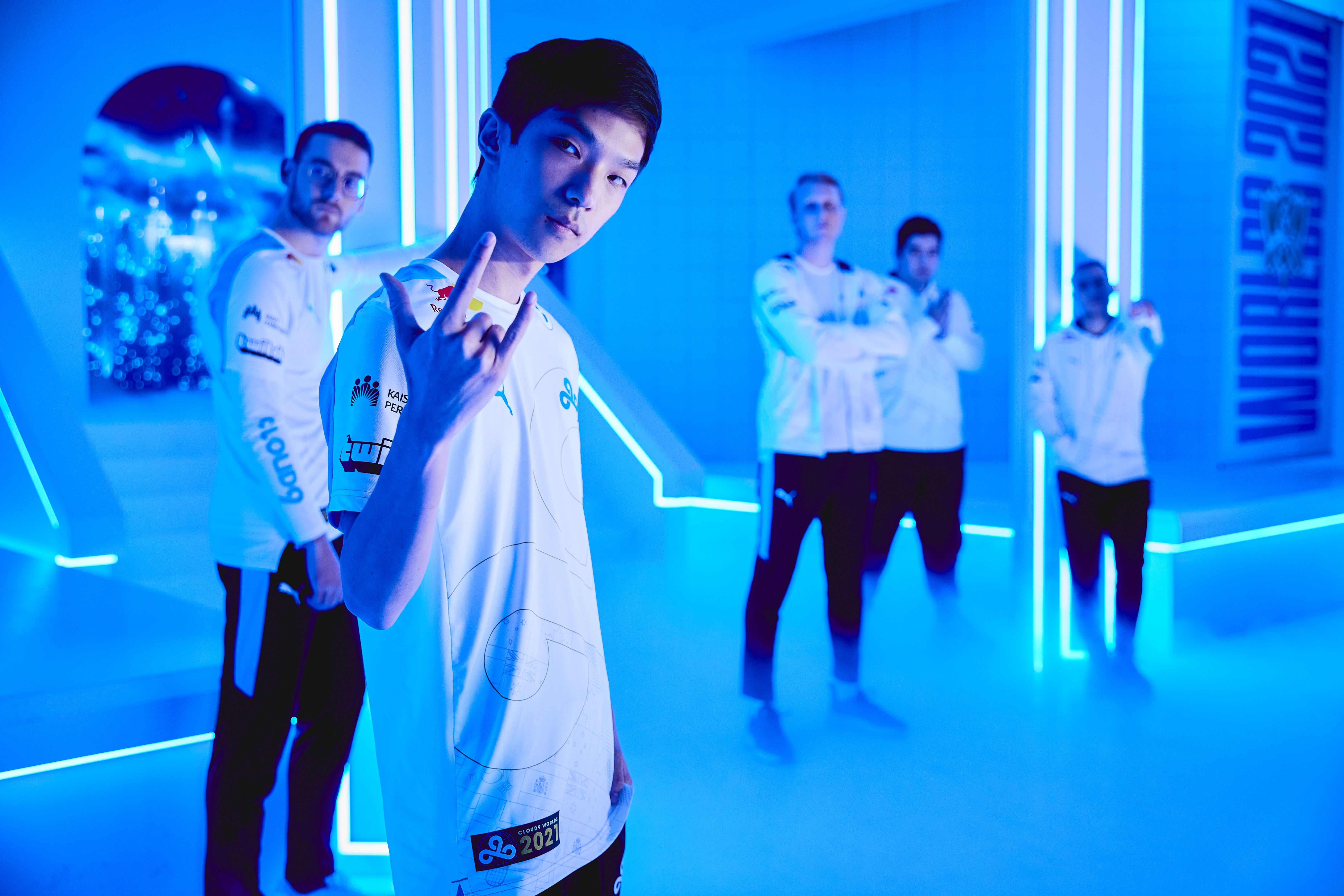 Cloud9 esports team in white jerseys on a blue neon-lit stage with fog. The foreground player makes a rock-on gesture, and a "Worlds 2021" sign is in the background.