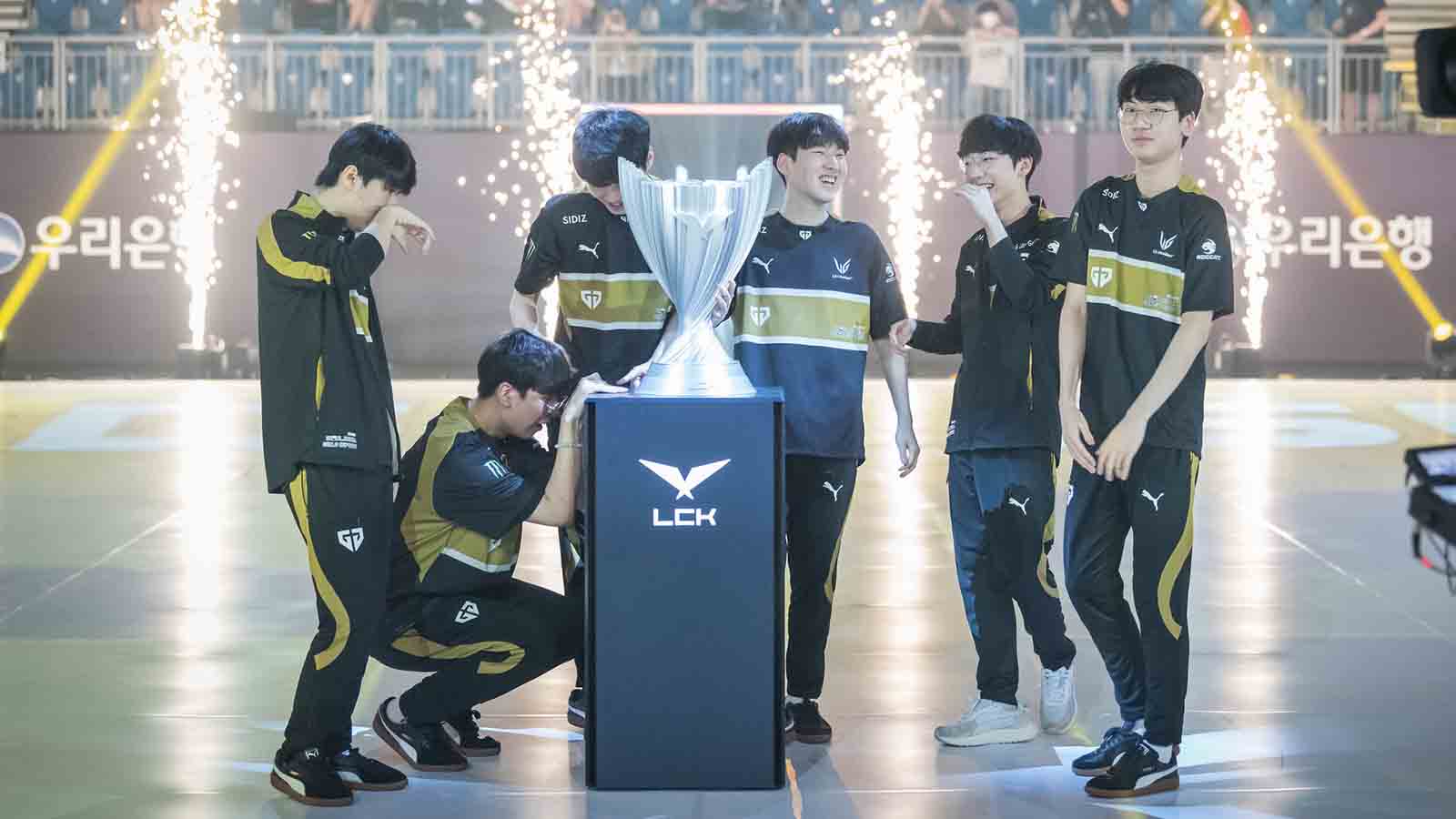 Six esports players in black and gold uniforms celebrate around a large silver LCK trophy with pyrotechnics in the background; some players are crying while others smile.
