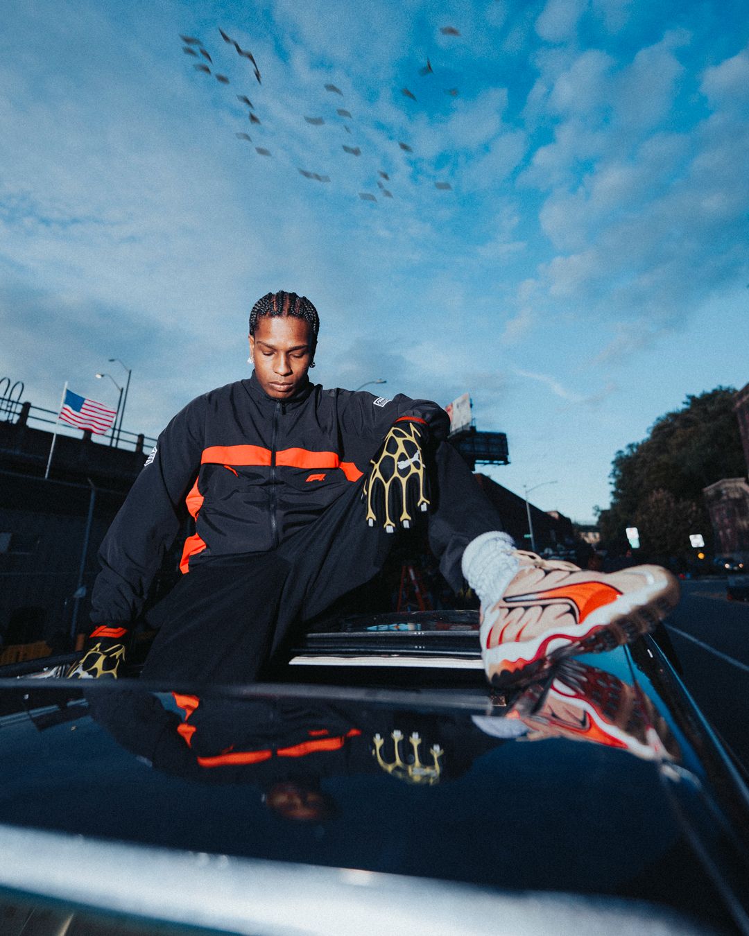 A man in a black and orange tracksuit and gold gloves sits on a car roof, reflected below him, under a cloudy sky with birds flying.