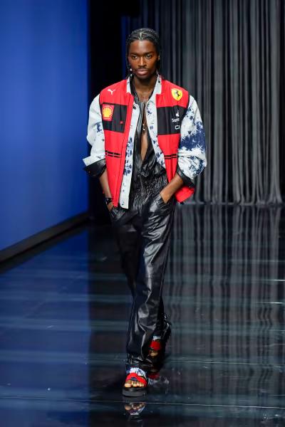 A male model with braided hair walks on a runway, wearing a red Ferrari racing jacket with tie-dye sleeves over a black leather outfit and colorful sandals.