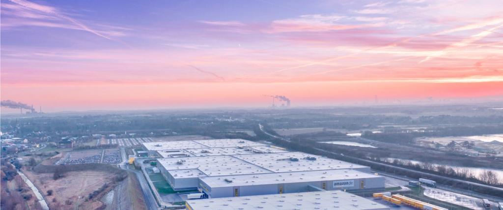 Aerial view of a large amazon.de fulfillment warehouse at pink sunrise with surrounding fields