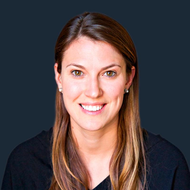Corporate headshot of a smiling woman with long light-brown hair in a black top on a dark blue background.