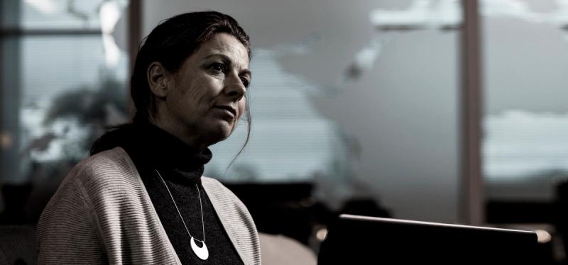 Woman in a turtleneck and cardigan looks ahead in an office with a blurred world map behind her