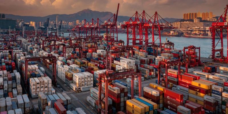 Aerial view of a vast container terminal with stacked Maersk containers and red cranes at sunset, city skyline behind