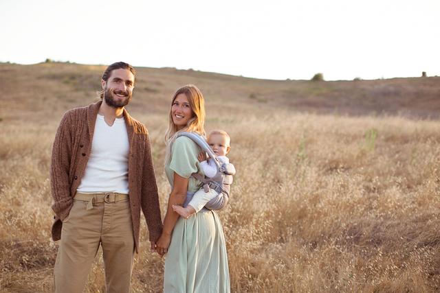 Couple standing in a golden field, mother carrying a baby in an Ergobaby carrier