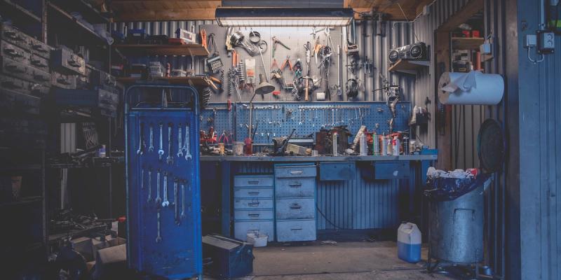 Dimly lit repair workshop with tools hung on a pegboard above a cluttered workbench and metal trash bin.