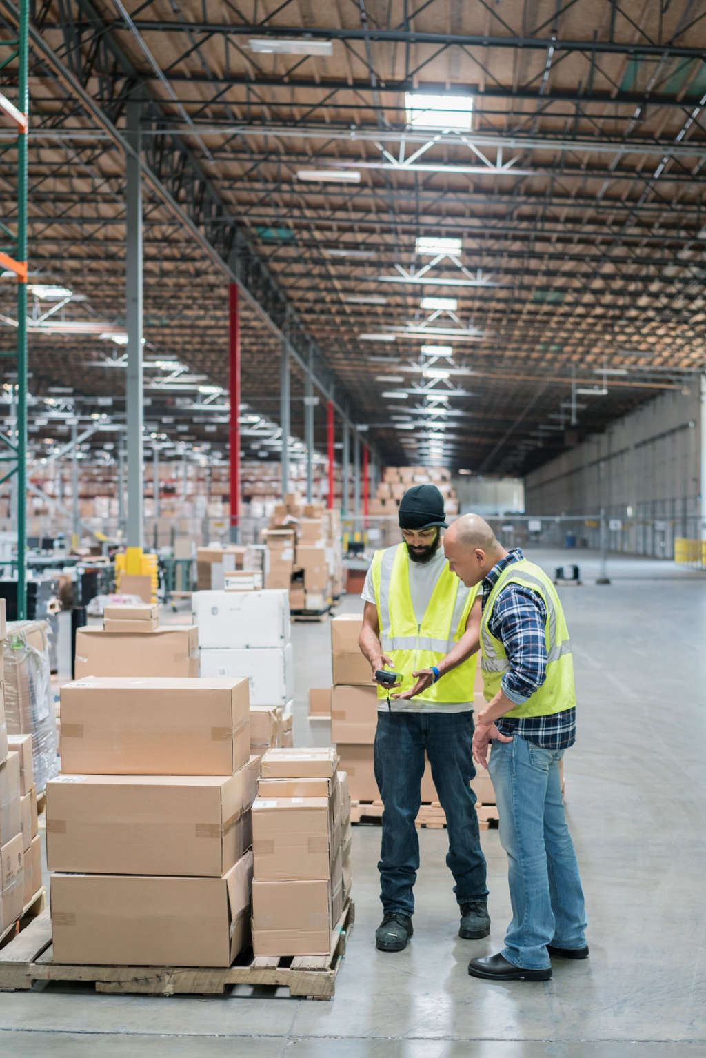 Two warehouse workers in safety vests examine inventory on a pallet in a large distribution center.