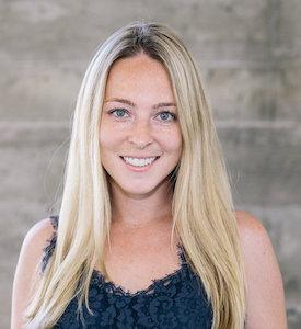 Headshot of a smiling blonde woman in a dark lace top against a concrete wall
