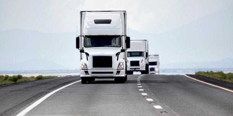 Convoy of white semi trucks driving toward viewer on an open desert highway with distant mountains