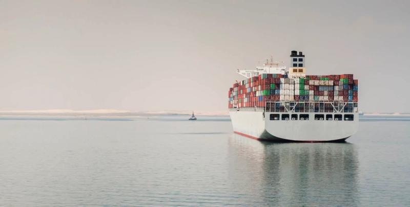 Container ship laden with stacked containers sailing through a canal flanked by desert banks