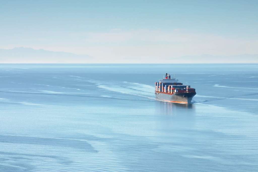 A cargo ship travels across calm open water with container stacks visible on deck.