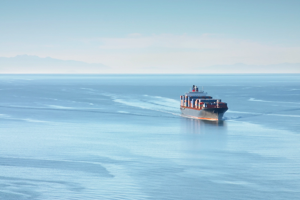 A cargo ship travels across calm open water with container stacks visible on deck.