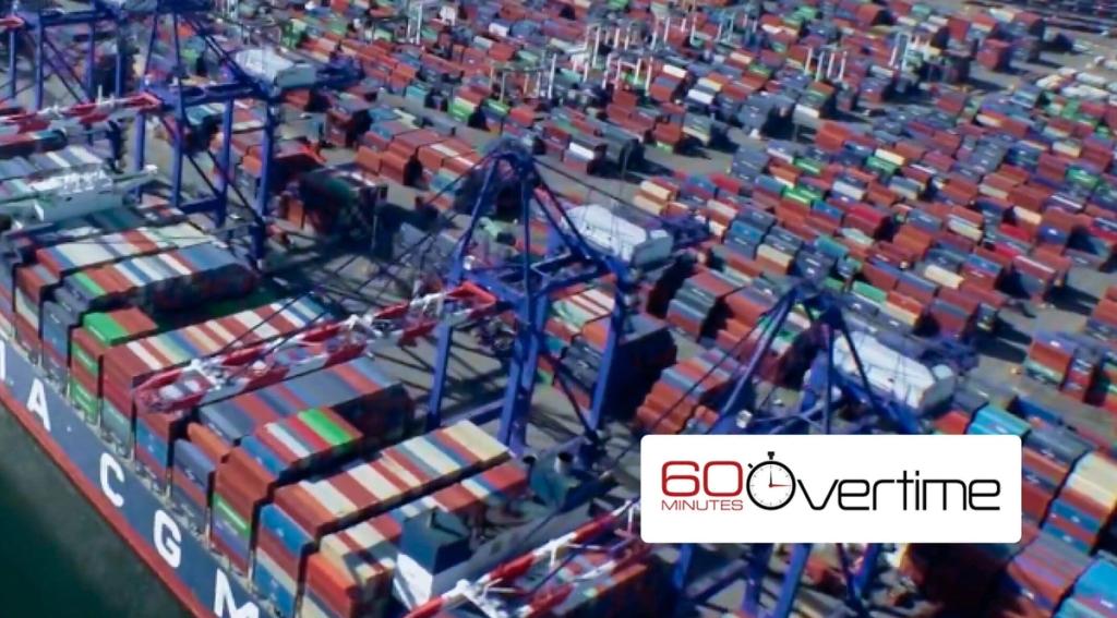 Aerial view of a container port packed with stacked shipping containers, ship cranes, and a CMA CGM vessel, with a 60 Minutes Overtime logo overlaid