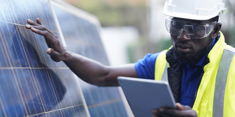 Worker in hard hat and safety vest inspects solar panels while holding a tablet outdoors