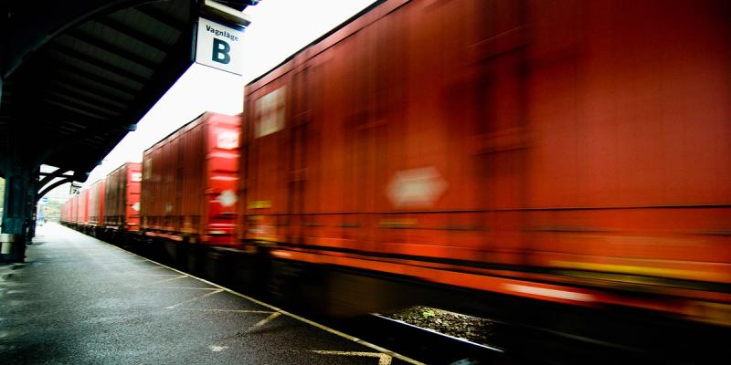 Red freight train rushing past a covered station platform marked Vagnlage B in motion blur