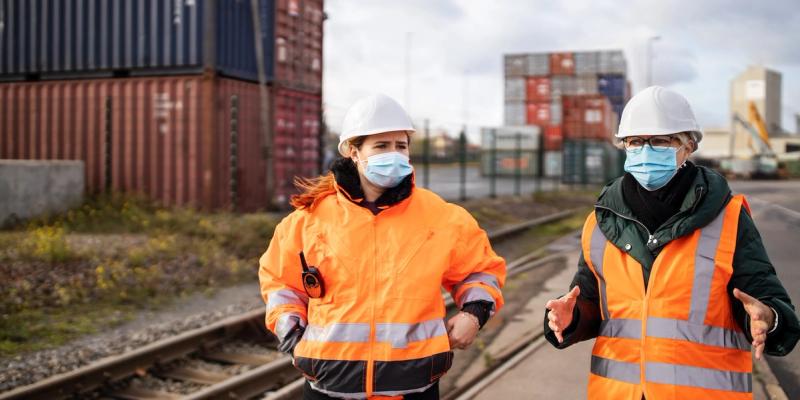 Two workers in hard hats, masks and hi-vis jackets walk along railway tracks past stacked shipping containers