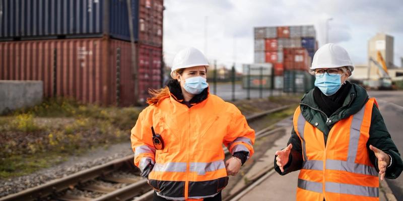 Two workers in hard hats, masks and hi-vis jackets walk along railway tracks past stacked shipping containers