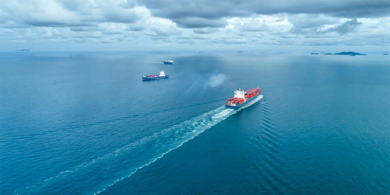 Aerial view of container ships crossing open blue ocean leaving long white wakes under cloudy sky