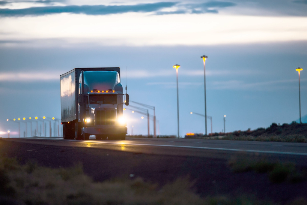 Semi-truck driving on a highway at dusk with headlights on and streetlights glowing.