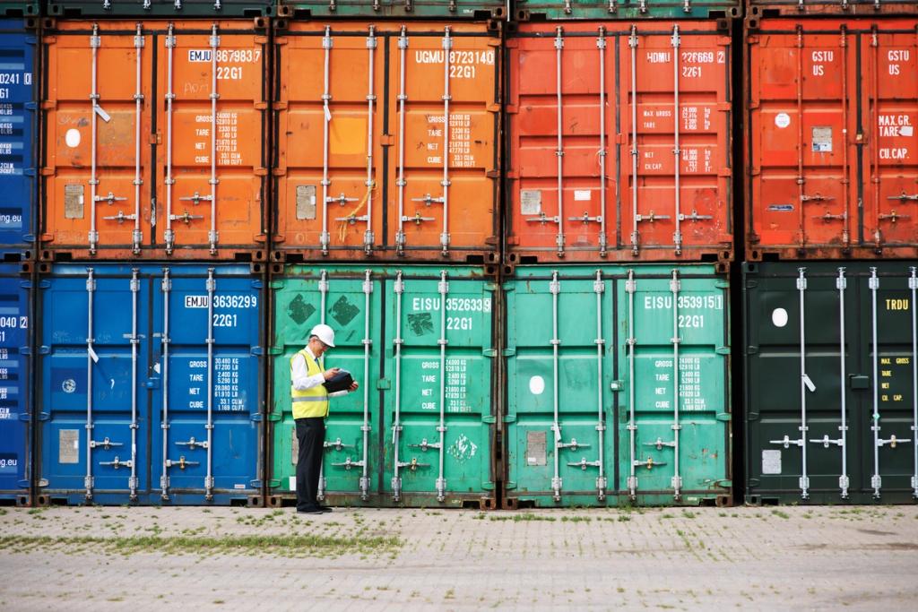 A worker in a hi-vis vest and hard hat stands writing on a clipboard in front of a stacked wall of colorful shipping containers at a port