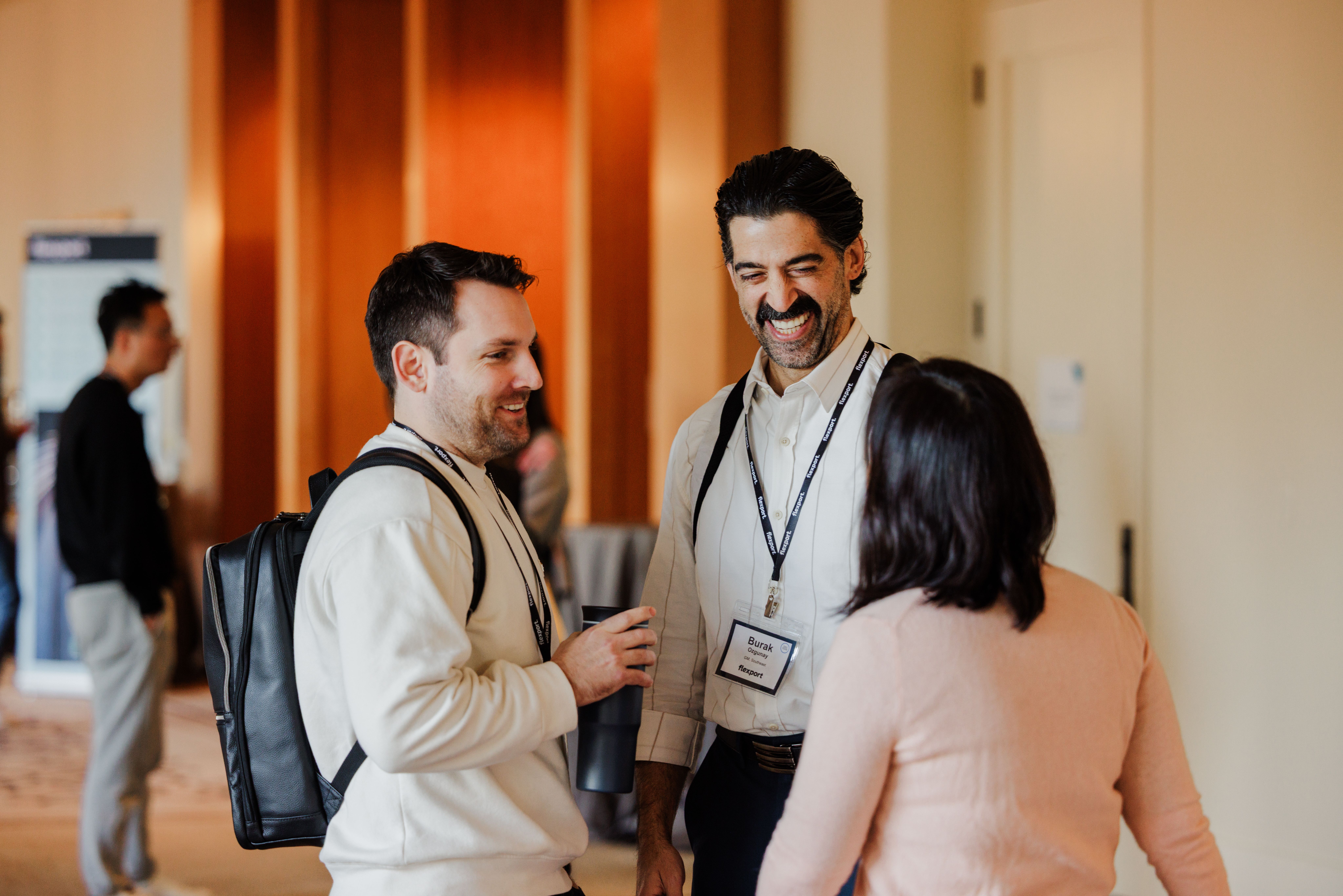 Three professionals with name badges converse in a hallway at a conference or networking event.
