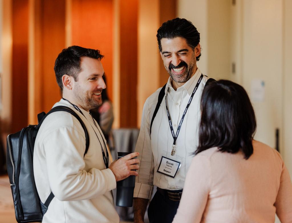 Three professionals with name badges converse in a hallway at a conference or networking event.