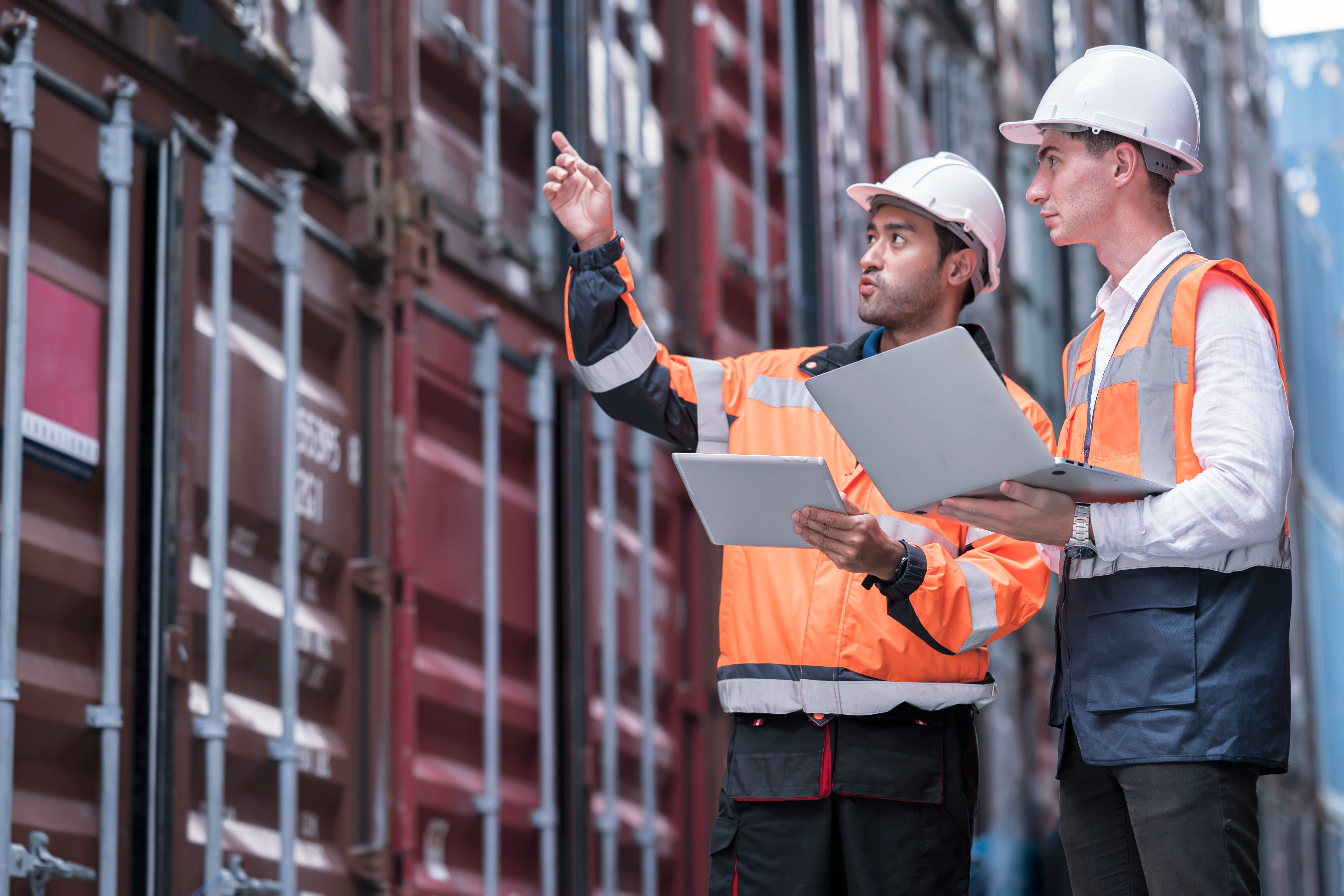 Two construction workers in safety vests and hard hats review plans while one points toward the building.