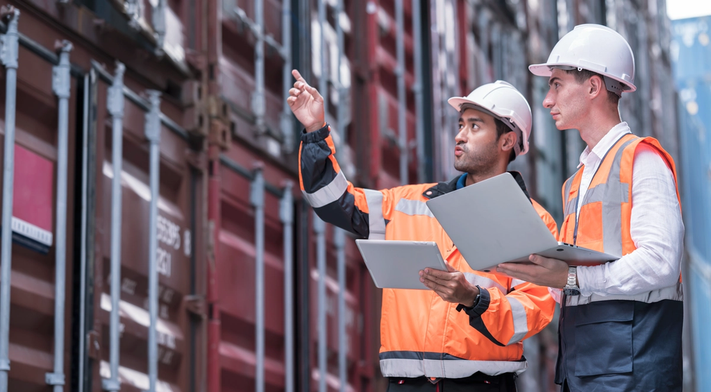 Two construction workers in safety vests and hard hats review plans while one points toward the building.