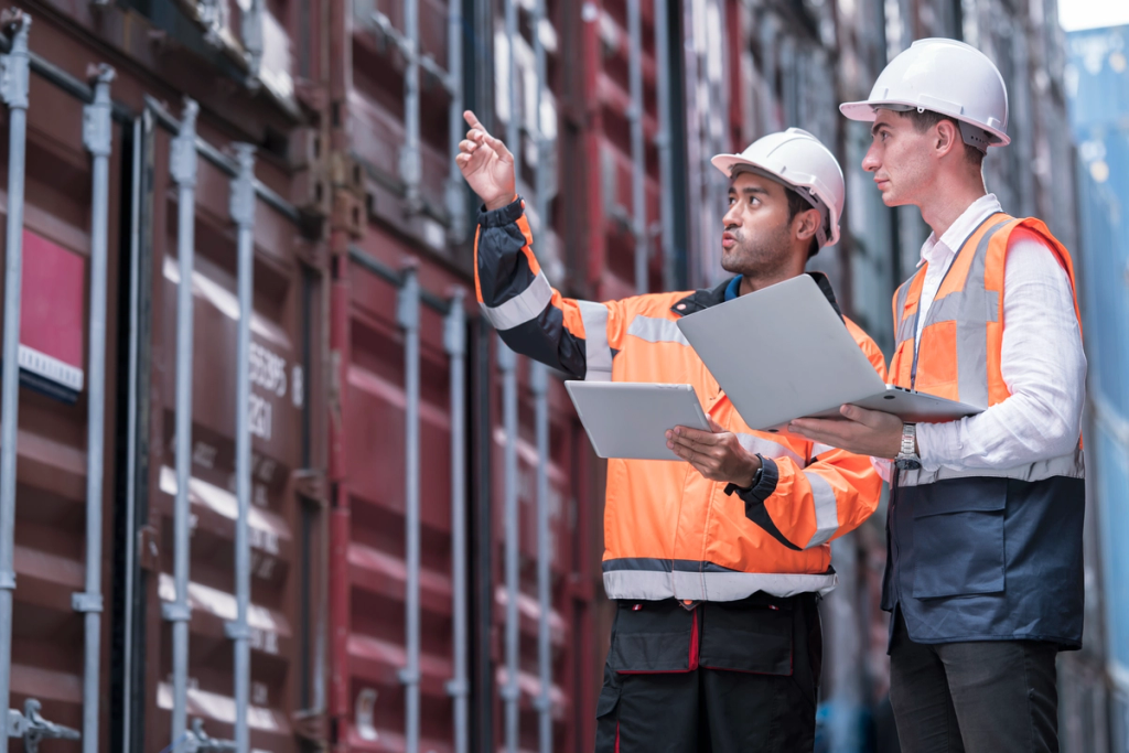 Two construction workers in safety vests and hard hats review plans while one points toward the building.