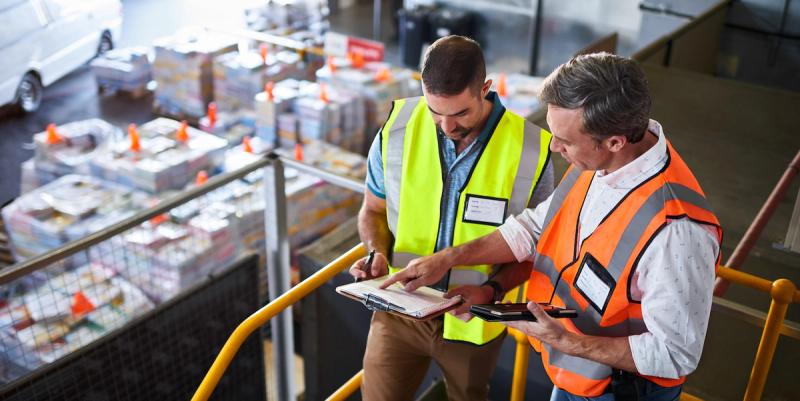 Two men in safety vests reviewing a binder on a mezzanine overlooking a warehouse floor
