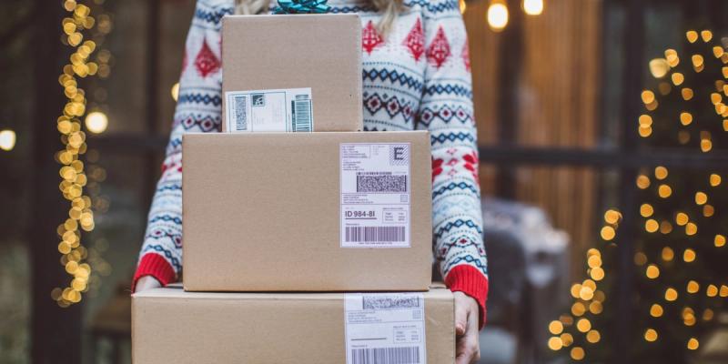 Person in a holiday sweater holding a stack of labeled cardboard shipping boxes with barcodes, framed by string lights