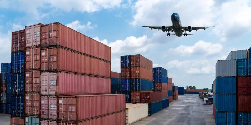 Passenger jet flying low over a container yard with stacked red and blue shipping containers