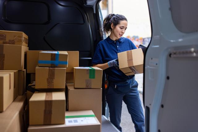 Delivery worker in blue uniform loading stacked cardboard parcels into the back of a white van