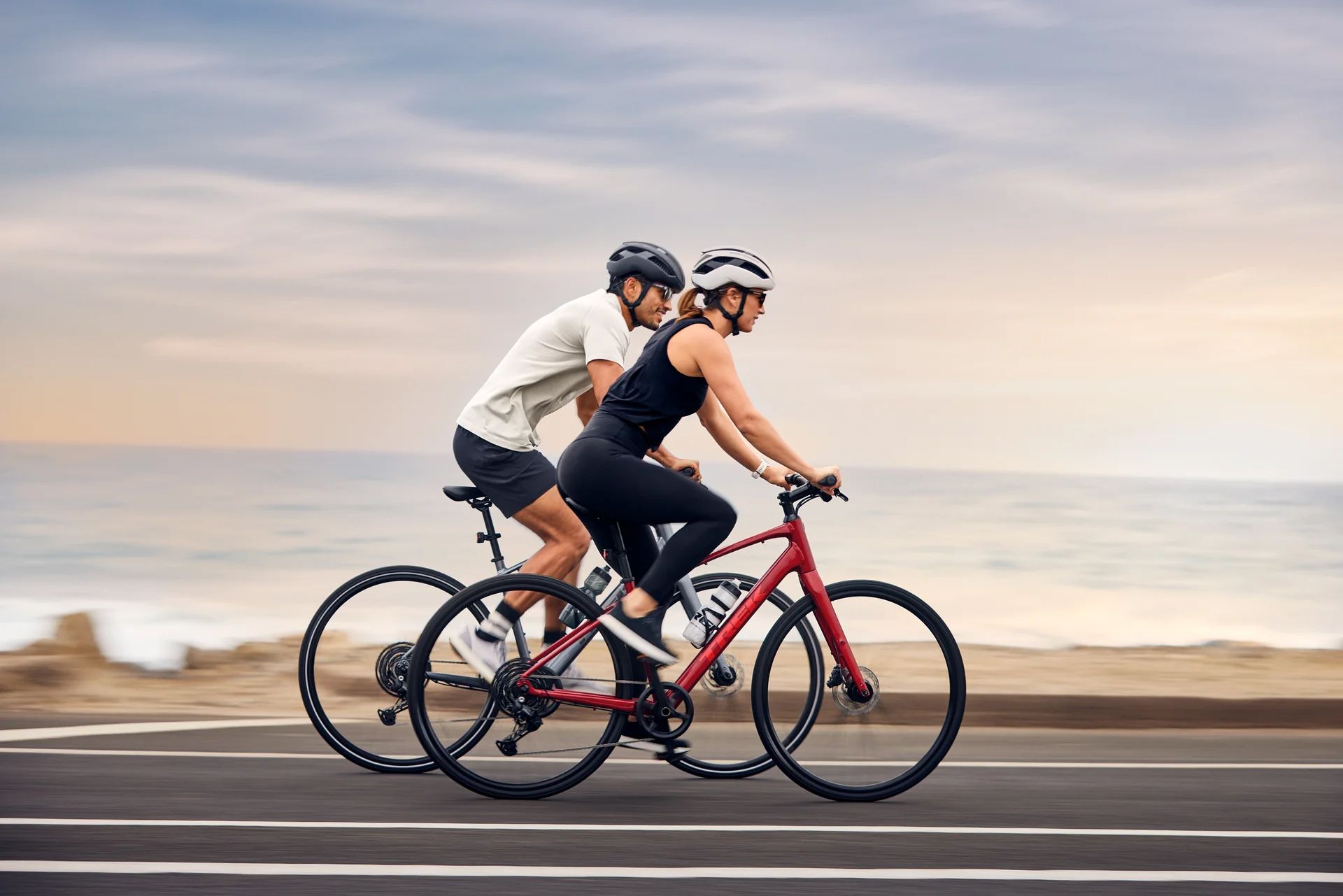 Two cyclists in helmets riding road bikes along a coastal road by the sea