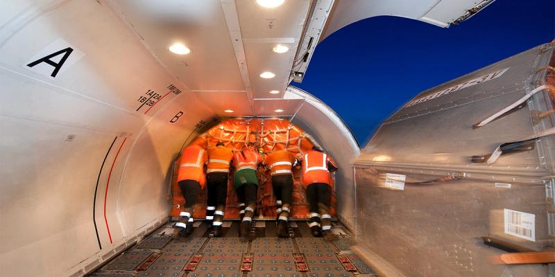 Five workers in orange hi-vis push a netted cargo pallet into the lit hold of a freighter aircraft at dusk.