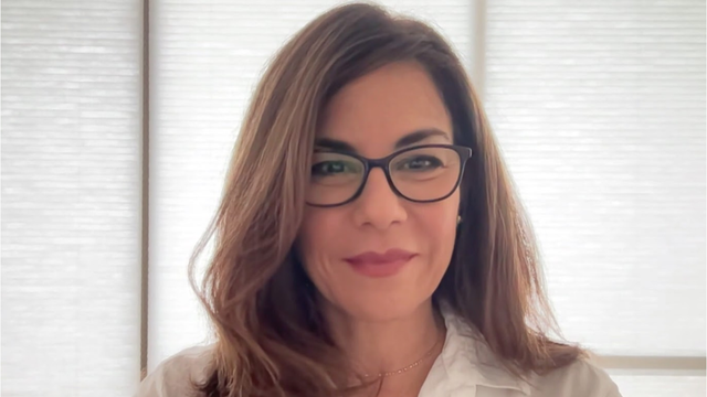 Headshot of a woman with long brown hair wearing dark-framed glasses and a white blouse in front of a window blind