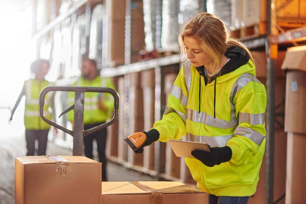 Warehouse worker in hi-vis jacket scanning a cardboard box with a handheld device, colleagues with pallet jack behind