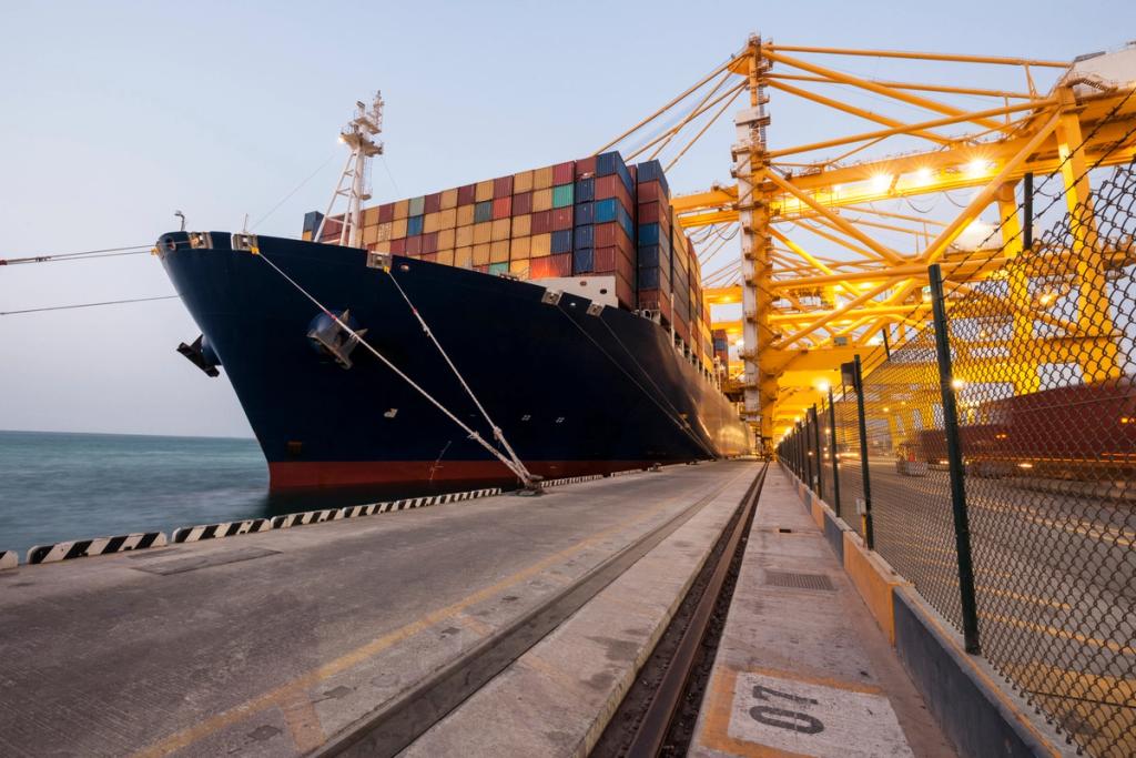 Container ship moored at a quay at dusk under yellow gantry cranes, bow at left, calm water and dockside rail track in foreground