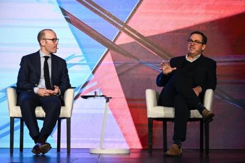 Two men in suits seated on a stage having a fireside chat with red and blue geometric backdrop